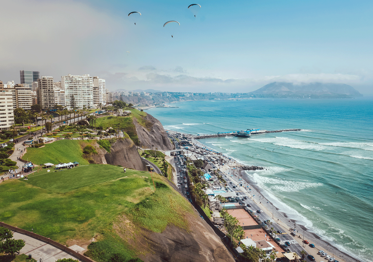 Dargestellt ist die Stadt Lima aus der Vogelperspektive. Die linke Seite der Fotografie zeigt die Stadt im Hintergrund und die begrünte Steilküste im Vordergrund, die rechte Seite das Meer.