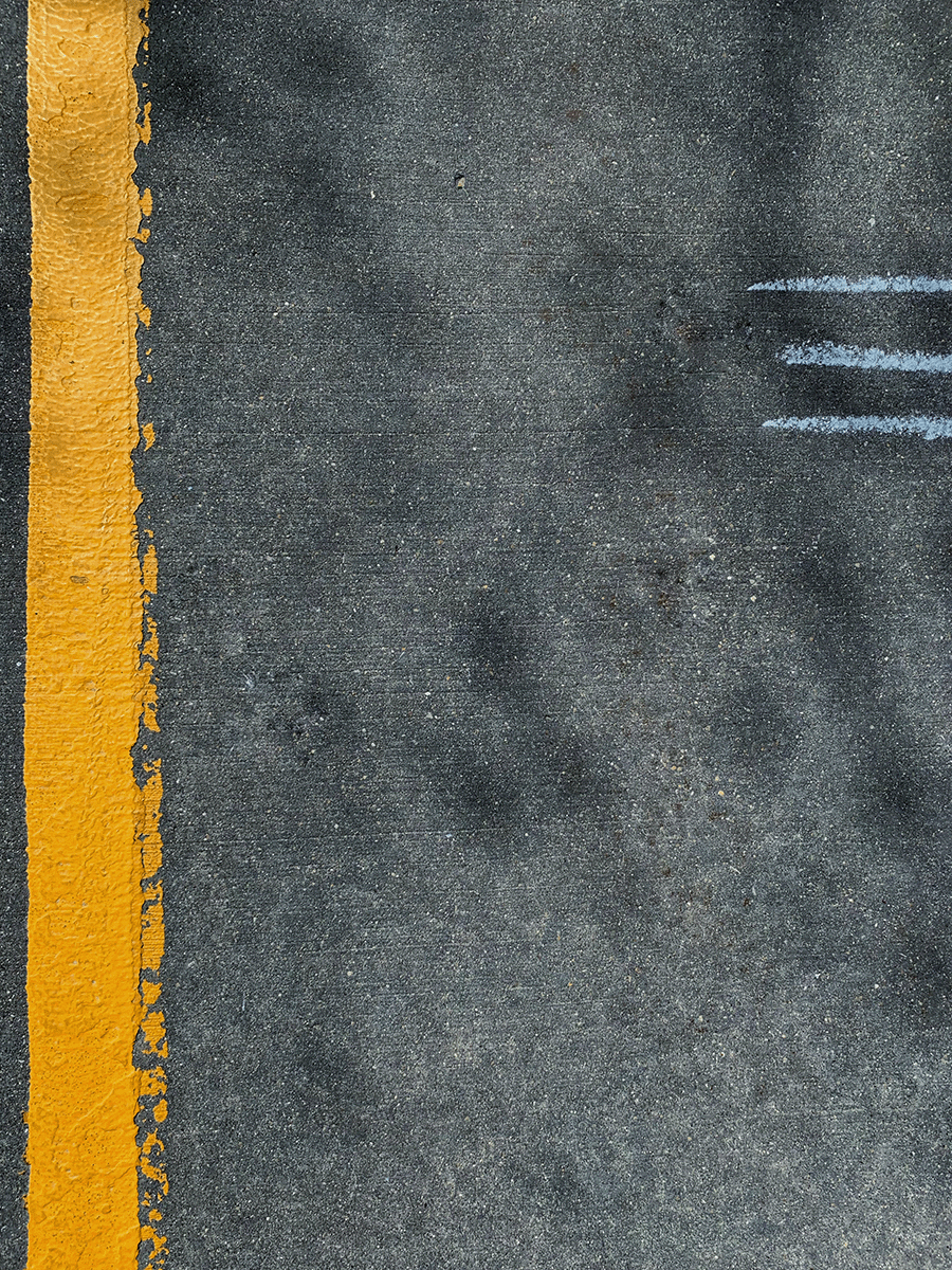 Das Bild zeigt eine Straßendecke aus der Vogelperspektive, auf der linken Seite ist ein gelber Fahrbahnstrefen auszumachen, der Rest ist eine graue Straße. Im rechten oberen drittel sind drei hellgraue Markierungen zu sehen.