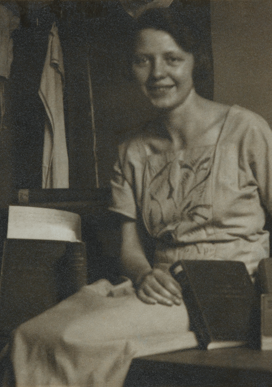 Auf dem Bild ist eine sitzende Frau in ihren zwanziger/ dreißiger Haaren mit zusammengebundenen Jahre zu sehen. Sie sitzt in einem Büro auf einem Schreibtisch, auf welchem sich zu ihrer linken Seite Bücher befinden.