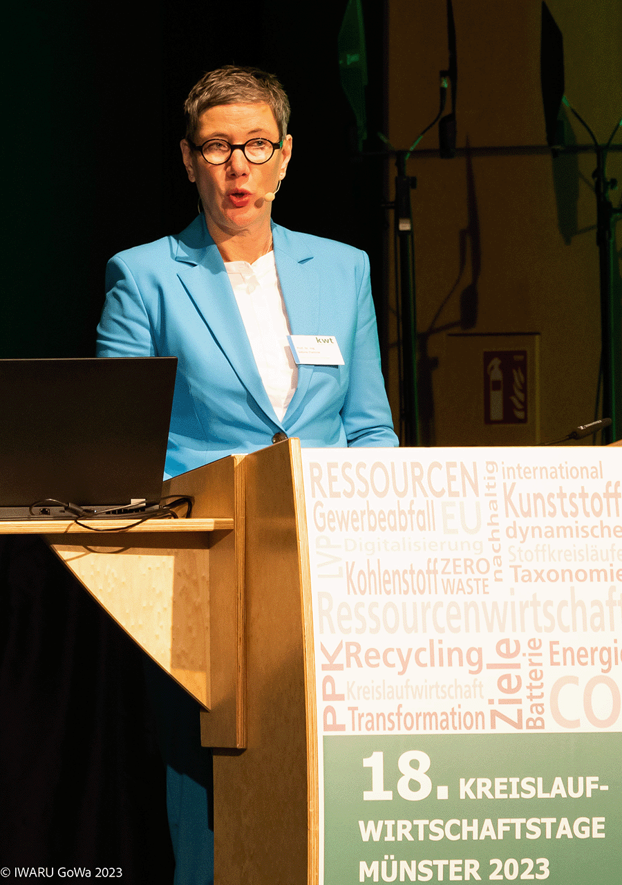 Auf dem Foto ist Frau Sabine Flamme hinter einem Rednerpult der Kreislaufwirtschaftstage Münster 2023 vor dunklem Hintergrund zu erkennen. Sie trägt ein weißes Oberteil sowie einen hellblauen Blazer.