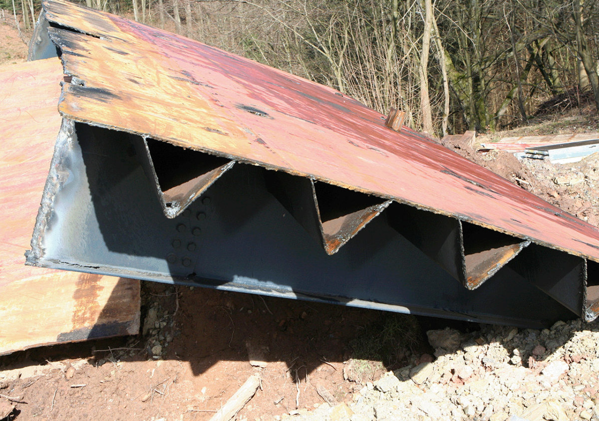Zu erkennen ist eine rötliche Stahlplatte, welche durch Dreiecke aus Stahl gestützt wird. An den Seiten befinden sich vertikale Stahlplatten und so liegt die Konstruktion auf einem Kiesuntergrund.
